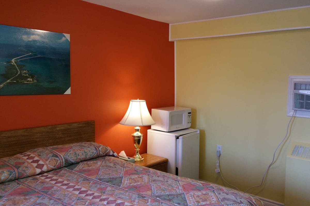 Hotellihuone, jossa on sänky, yöpöytä lampun kanssa, mikroaaltouuni ja seinälle asennettu ilmastointilaite.