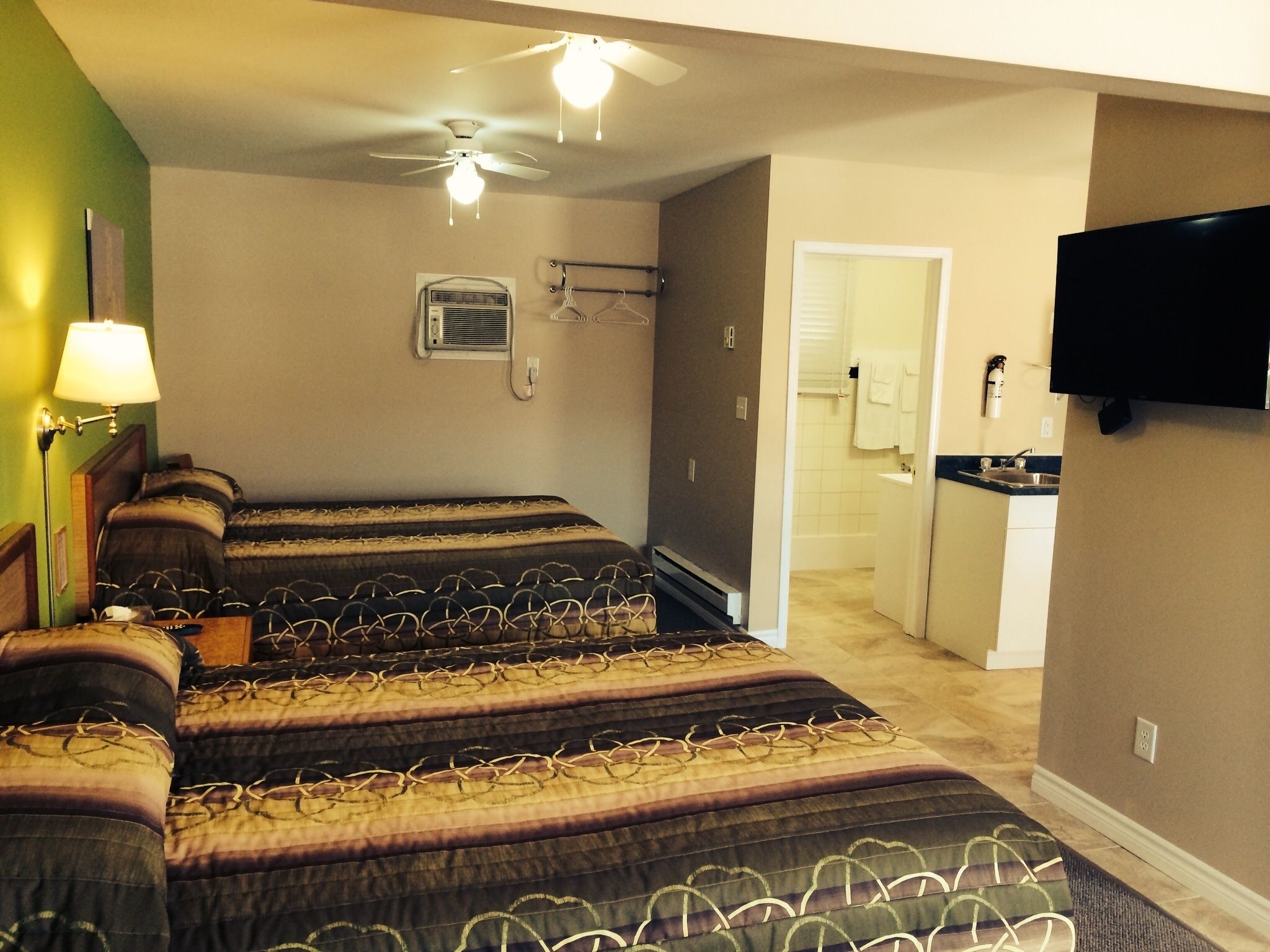 A hotel room with two beds, a ceiling fan, a TV, a small kitchenette, and a bathroom visible through an open door.