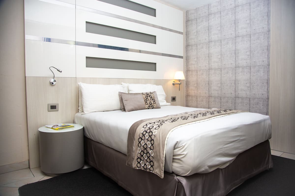 A modern hotel room with a large bed, a bedside table, a wall-mounted lamp, and a small round trash bin.