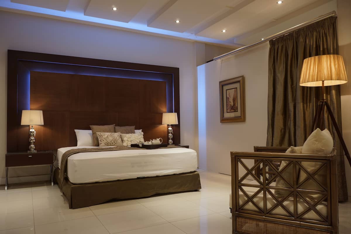 A modern hotel room with a large bed, two bedside tables with lamps, a nightstand, a decorative chair, and a large framed picture on the wall.