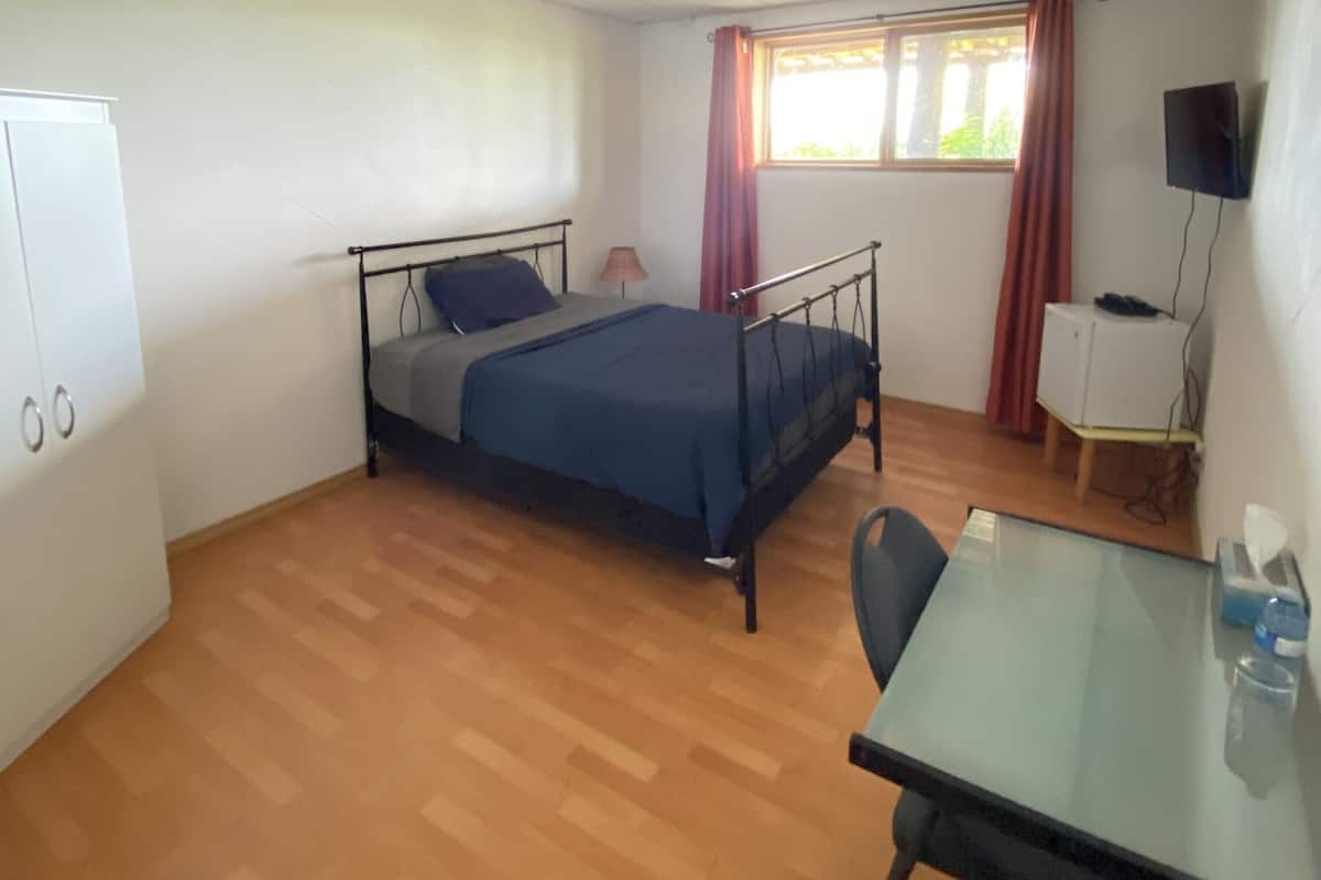 Ein Einzelbett mit Metallgestell, ein Nachttisch, ein Stuhl, ein Schreibtisch, ein Fernseher auf einem Schrank und ein Fenster mit Vorhängen.