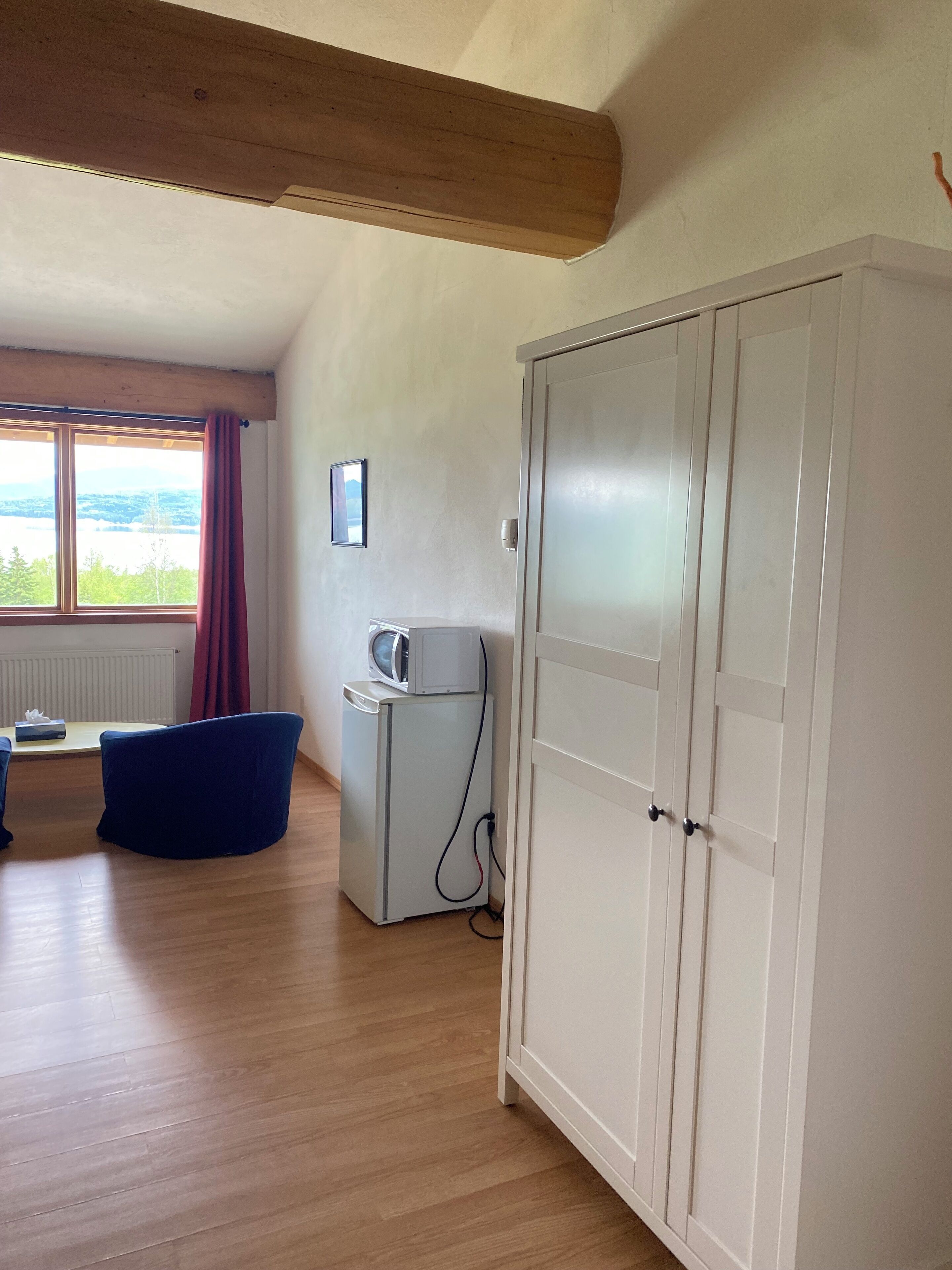 Una habitación con un armario blanco grande, un refrigerador blanco pequeño, una silla azul, una mesa de madera y una ventana con vistas al exterior.