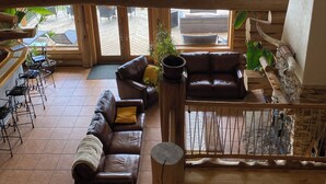 A spacious loft-style room with a high ceiling, large windows offering a view of the outdoors, a seating area with leather sofas and a coffee table, and a wooden accent wall. - Williston Lake Resort (Hudson's Hope)