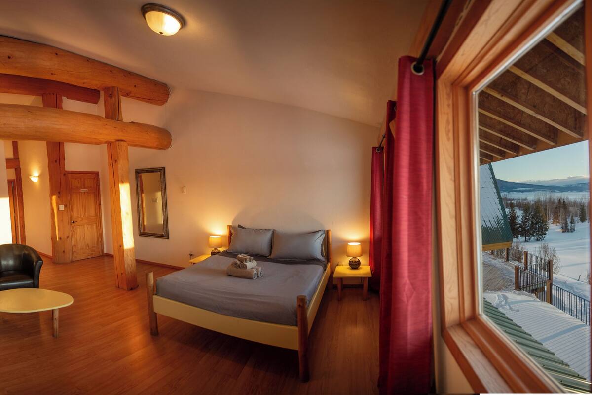 Ein Schlafzimmer mit einem Bett, zwei Stühlen, einem kleinen Tisch, Holzbalken, einem Fenster mit Blick auf eine verschneite Landschaft und einer Tür.