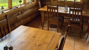 A wooden cabin interior with a dining table and chairs, large windows with a view of greenery and mountains, and a hanging light fixture. - Williston Lake Resort (Hudson's Hope)