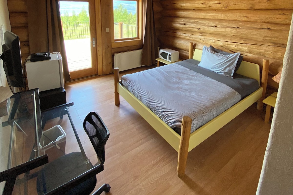Ein Schlafzimmer in einer Blockhütte mit Bett, Schreibtisch, Stuhl und Blick ins Freie.