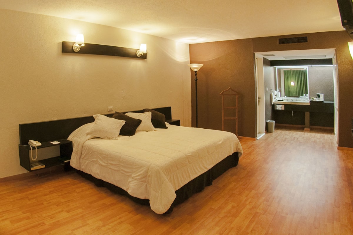 A hotel room with a bed, bedside table, lamp, and a bathroom visible through an open door.