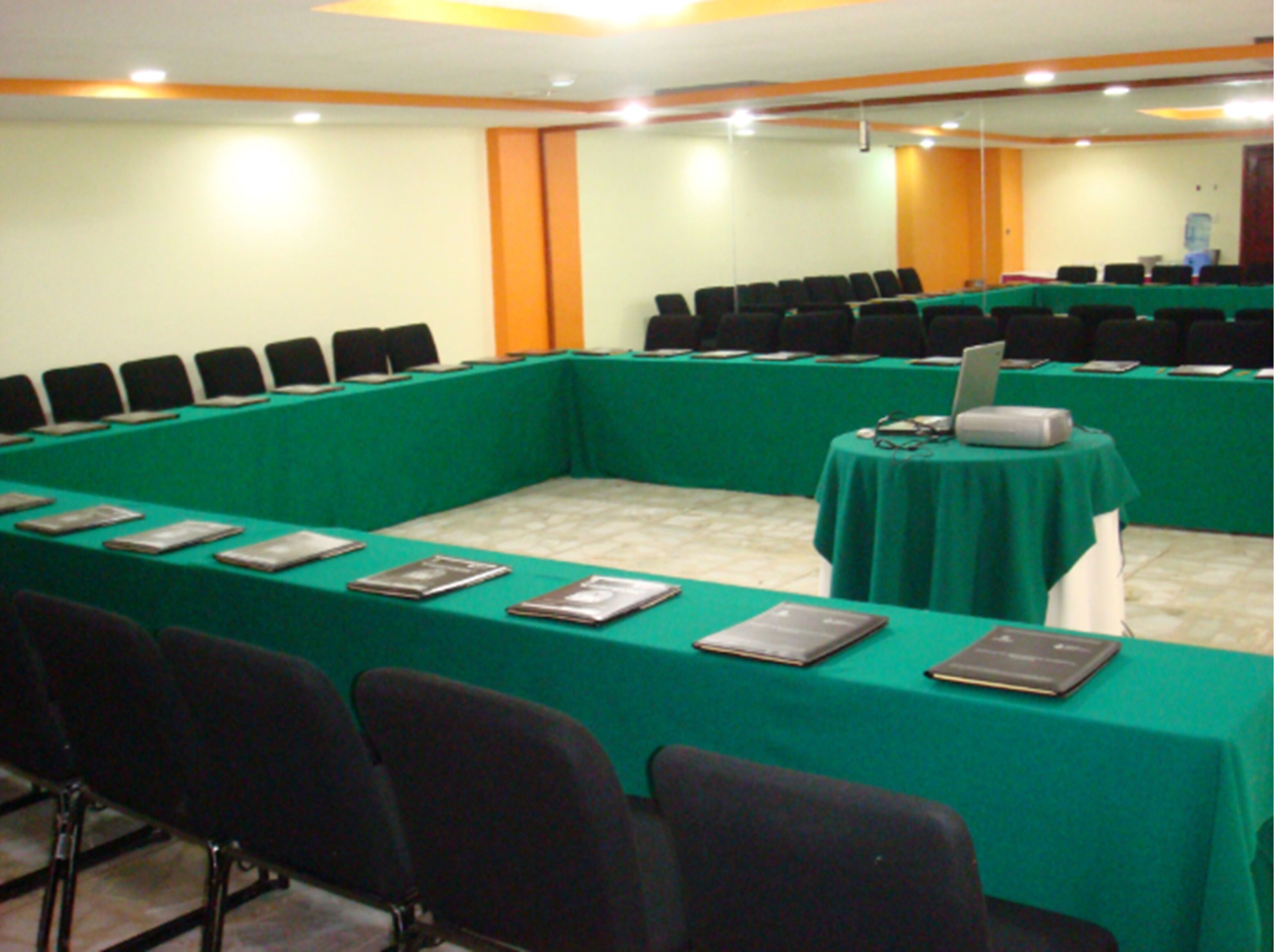 Una sala conferenze con un lungo tavolo ricoperto da una tovaglia verde, diverse sedie e un laptop su un tavolino.