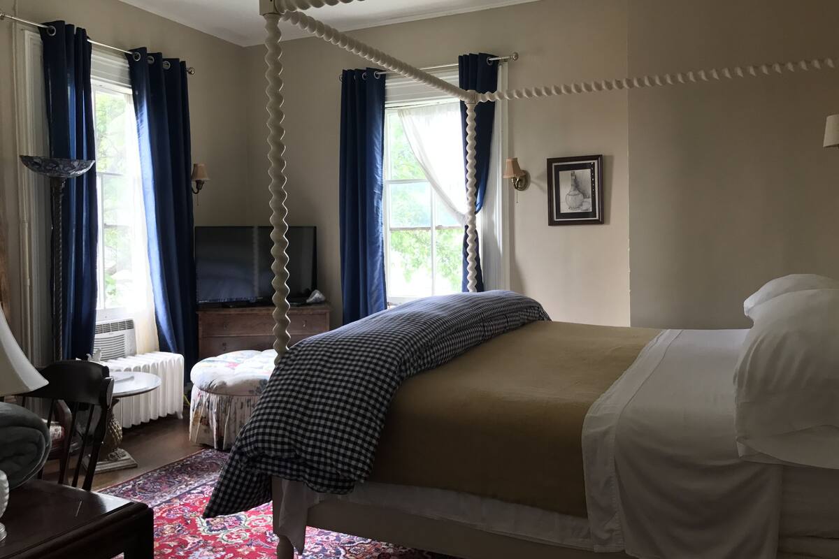 A four-poster bed with a checkered bedspread, a white canopy, and a red patterned rug.