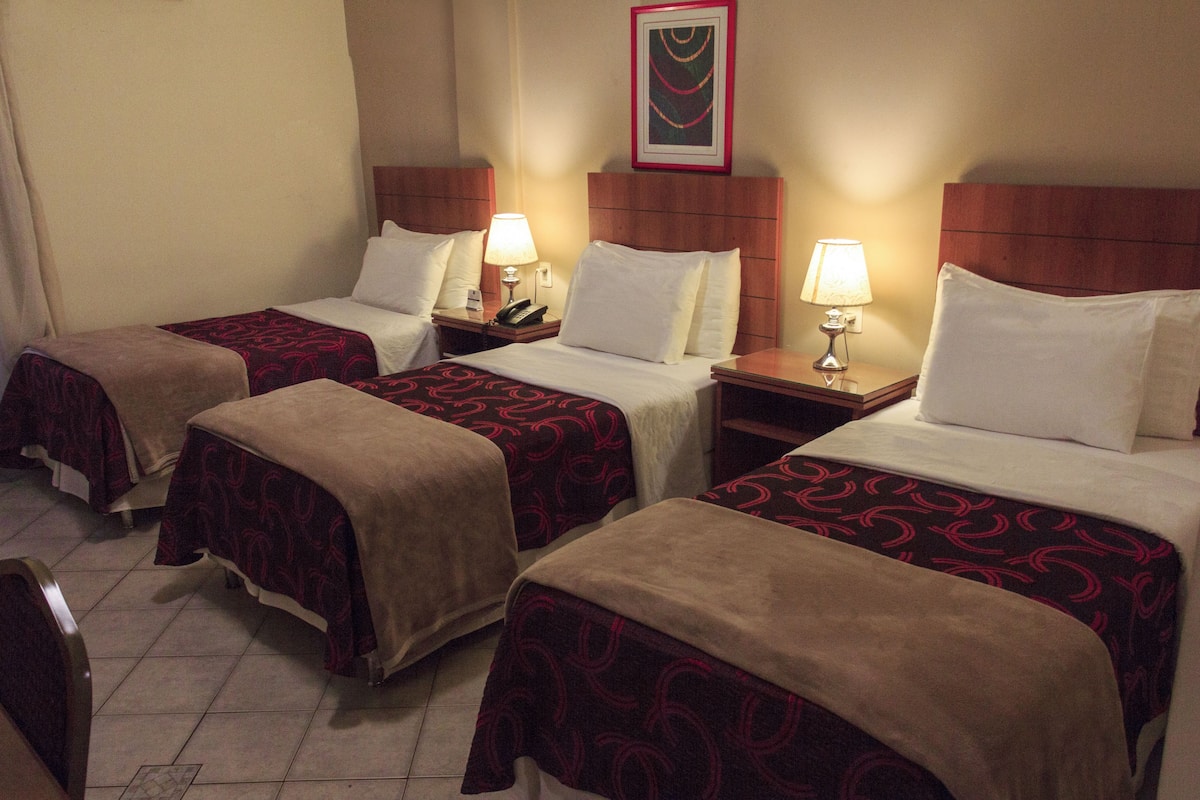 A hotel room with three single beds, each with a wooden headboard, bedside tables with lamps, and a framed artwork above the beds.