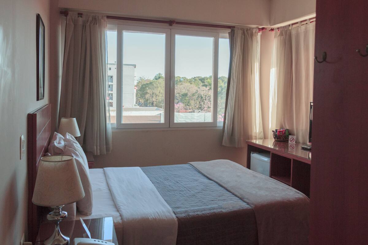 A hotel room with a large window, a bed, a desk with a lamp, and a small table with a basket.