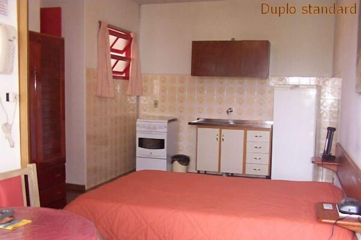 A compact room with a bed, a small table, a kitchenette, and a window with curtains.