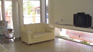 A living room with a beige sofa, a television on a stand, and a view of the outside through a glass door. - Residencial Sol e Mar de Floripa (Florianópolis)