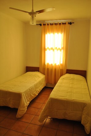 Una habitación pequeña con dos camas individuales, un ventilador de techo y una ventana con cortinas amarillas.