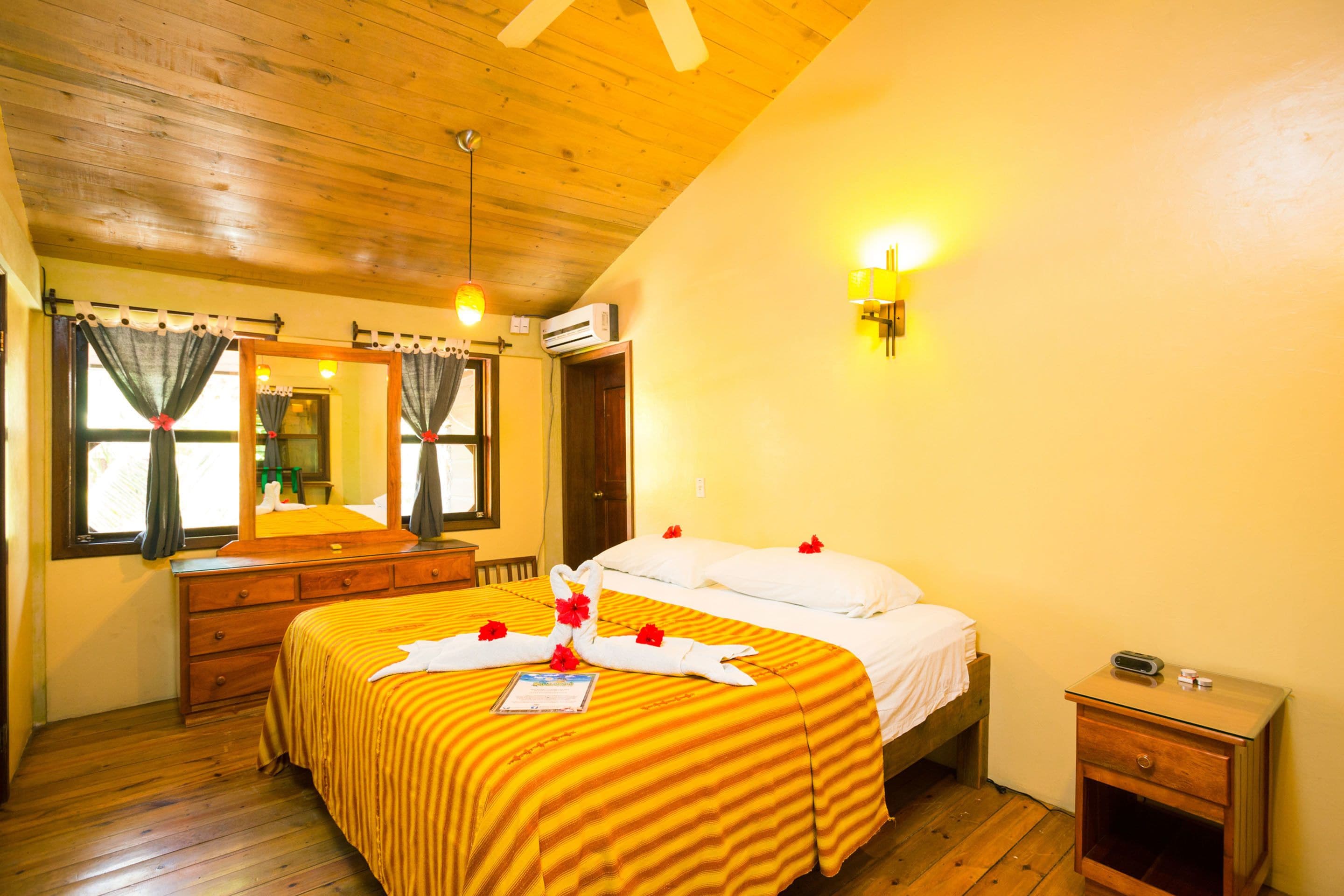 Habitación de hotel con techo de madera, cama con colcha a rayas, mesita de noche con radio y espejo sobre el tocador.