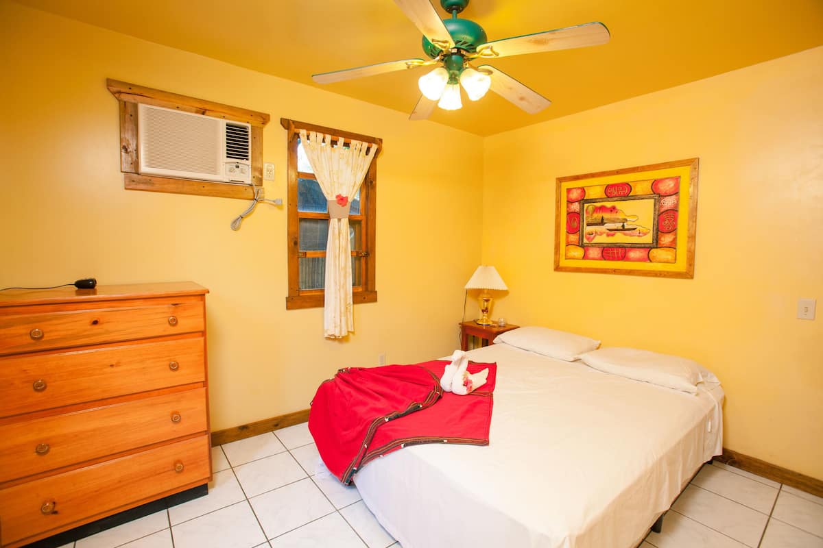 Un dormitorio con cama, cómoda, ventilador de techo, ventana con cortinas y aire acondicionado instalado en la pared.