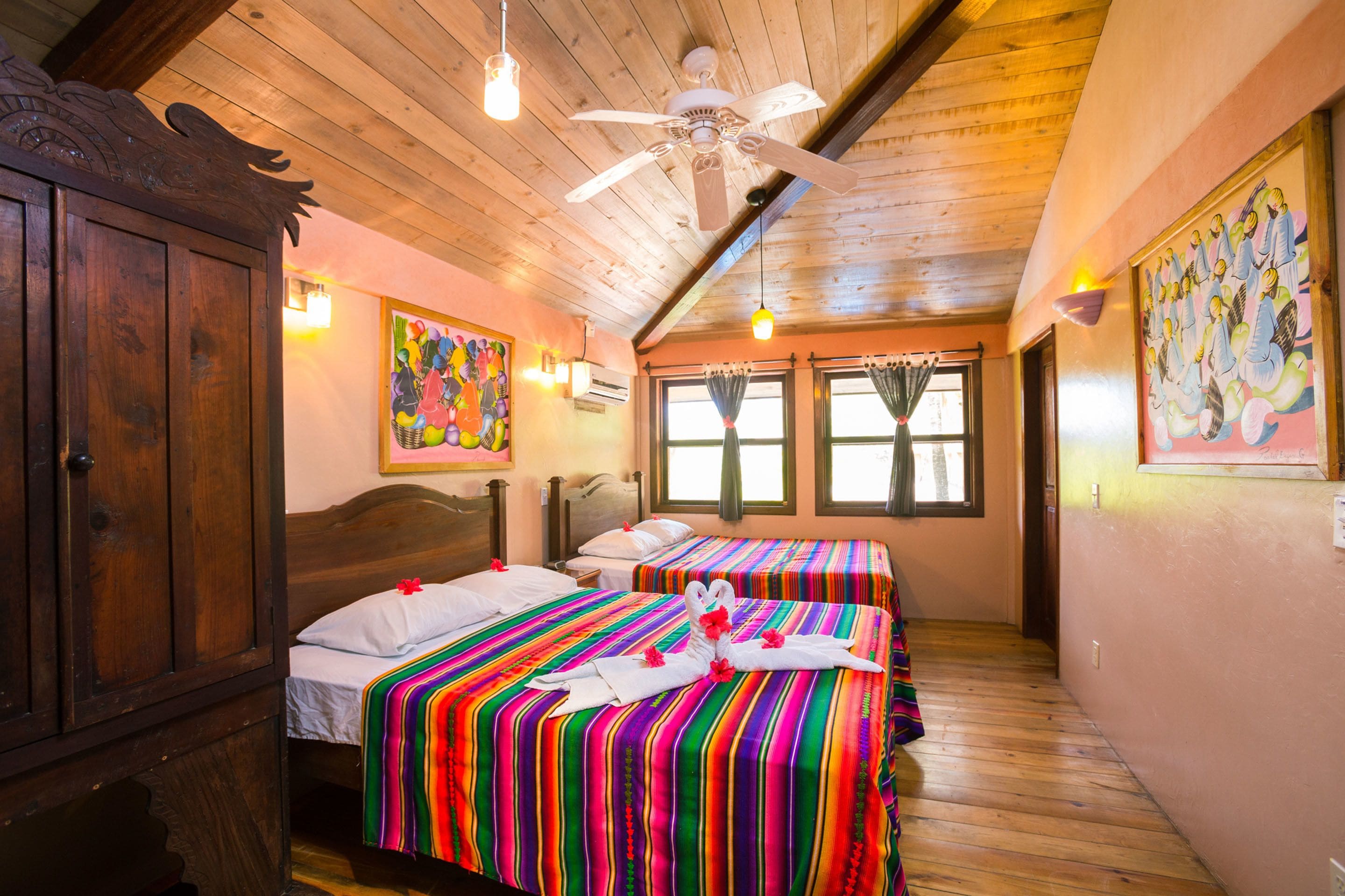 A room with two beds, wooden ceiling beams, a ceiling fan, and colorful artwork on the walls.