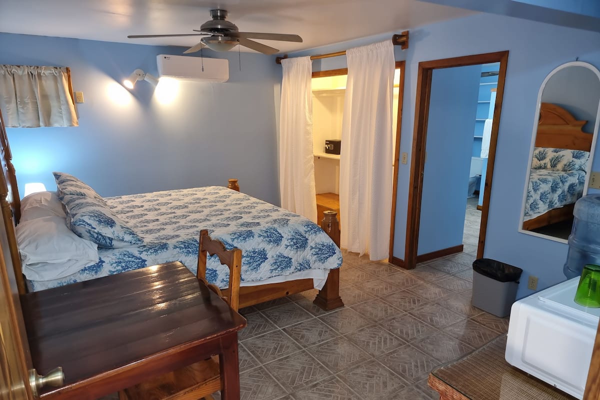 Un dormitorio con una cama, una silla de madera, un escritorio, un ventilador de techo y un dispensador de agua.