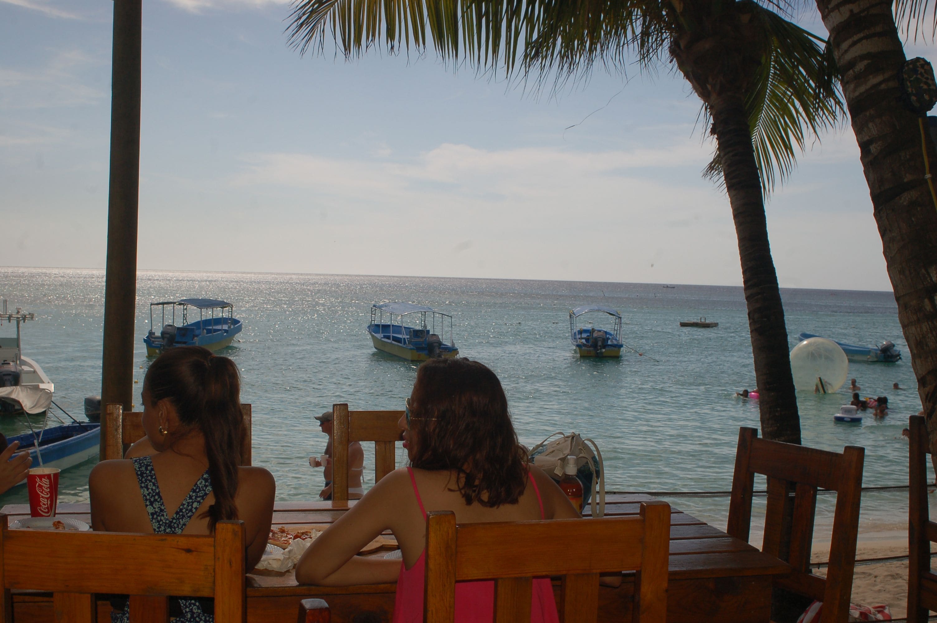 A wooden table and chairs at a beachside restaurant with a view of the sea and boats.