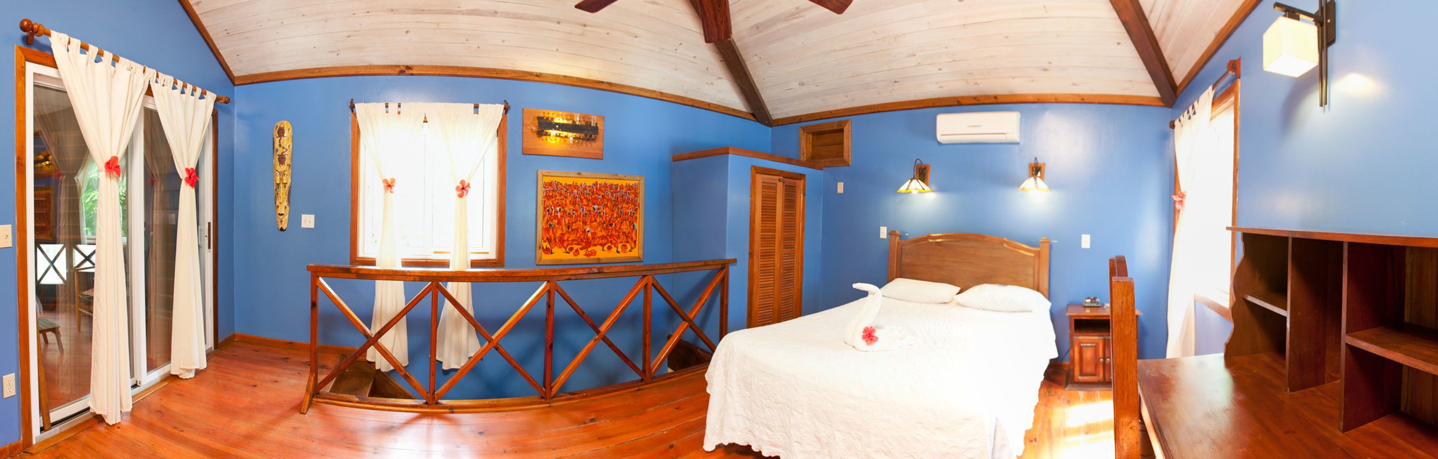 Una habitación con una cama, una mesa de madera, un armario pequeño y un ventanal con cortinas.