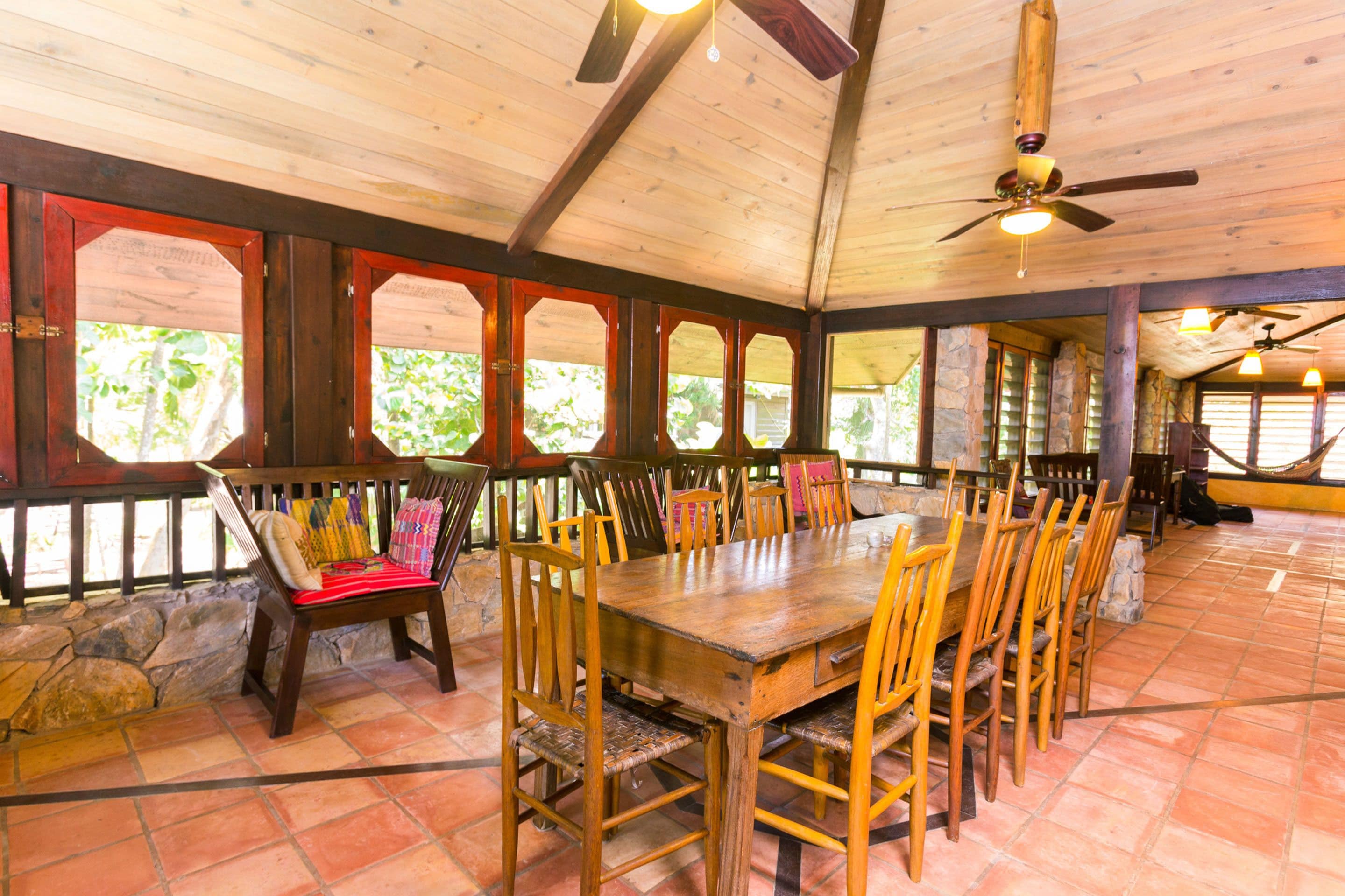 A spacious dining area with a large wooden table and chairs, stone walls, and a high ceiling with wooden beams.