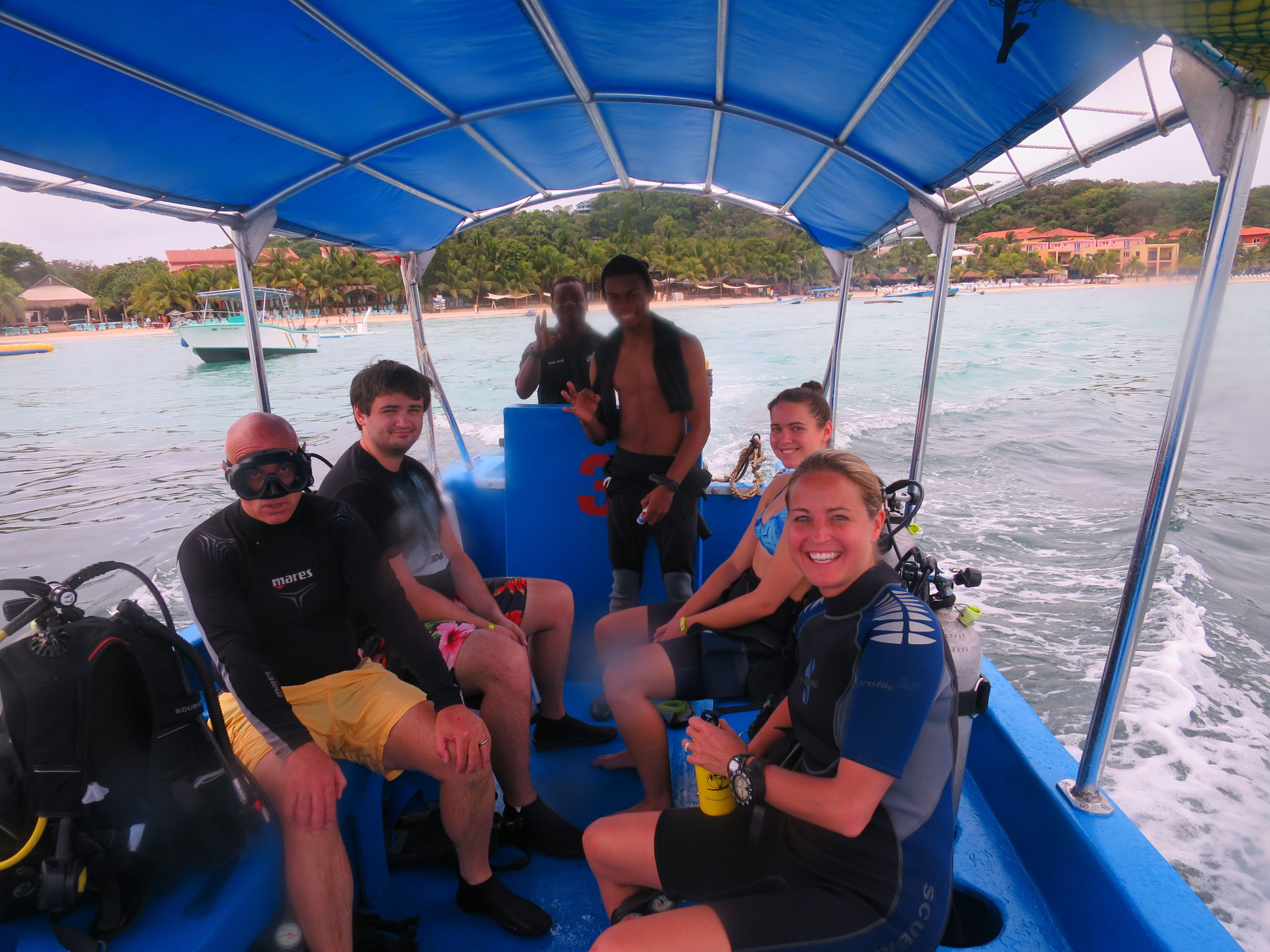 A group of people on a boat with a blue canopy, some wearing diving gear, and a beach visible in the background.