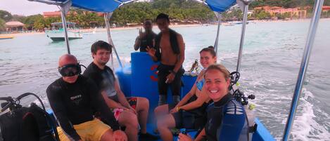 A group of people on a boat with a blue canopy, some wearing diving gear, and a beach visible in the background.