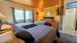 A hotel room with two beds, a wooden ceiling, a window with curtains, a bedside table with a lamp, and a framed picture on the wall. - The Jay Village Inn (North Troy)