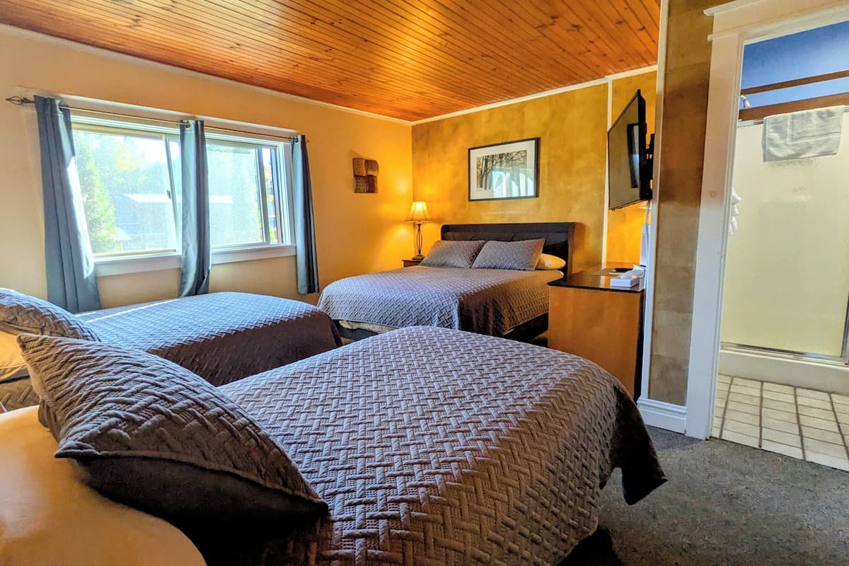 A hotel room with two beds, a wooden ceiling, a window with curtains, a bedside table with a lamp, and a framed picture on the wall.
