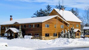 A large wooden lodge with multiple windows and a snow-covered roof, surrounded by snow and trees. - The Jay Village Inn (North Troy)