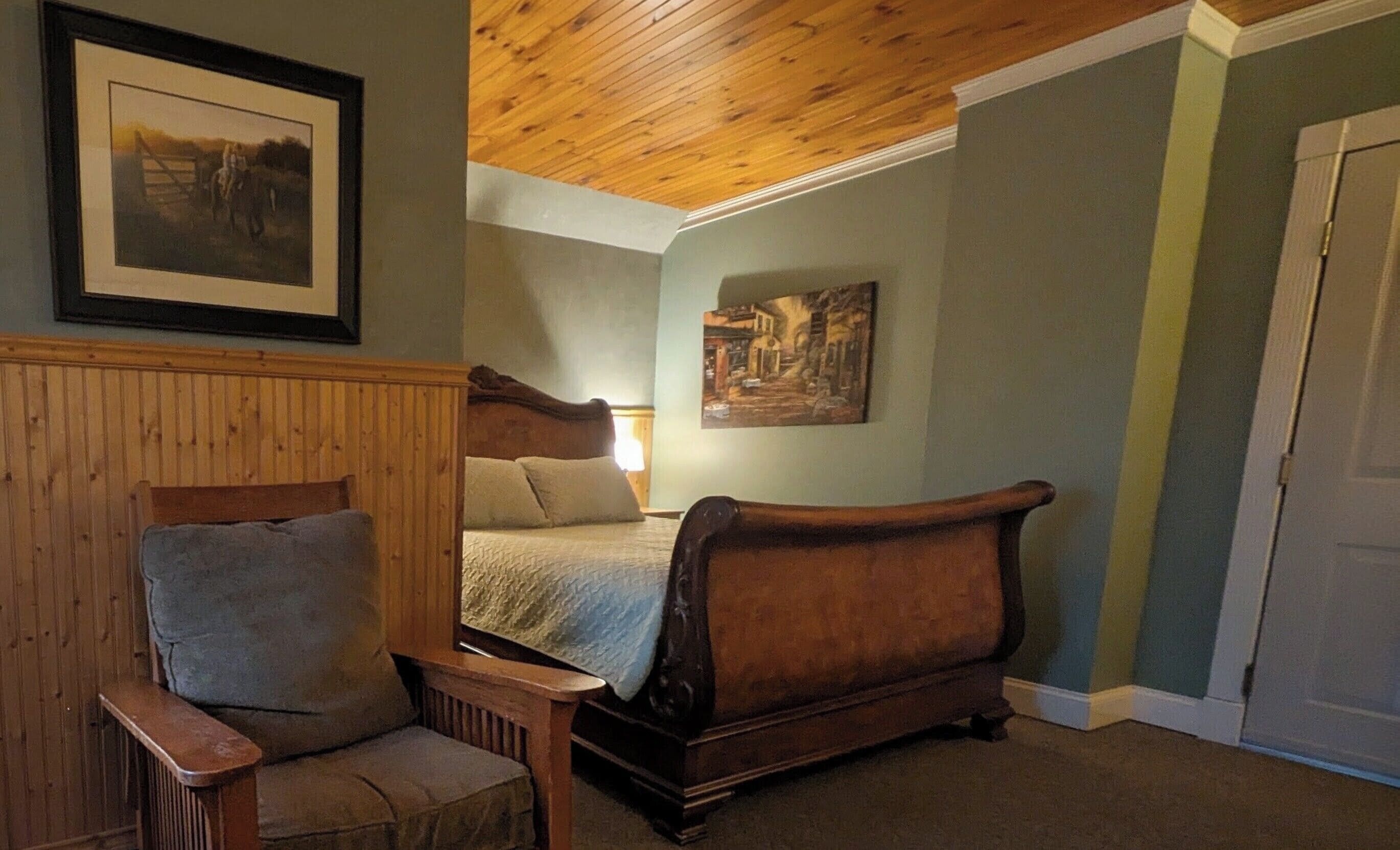 A room with a wooden ceiling, a bed with a headboard, a chair, and two framed pictures on the wall.