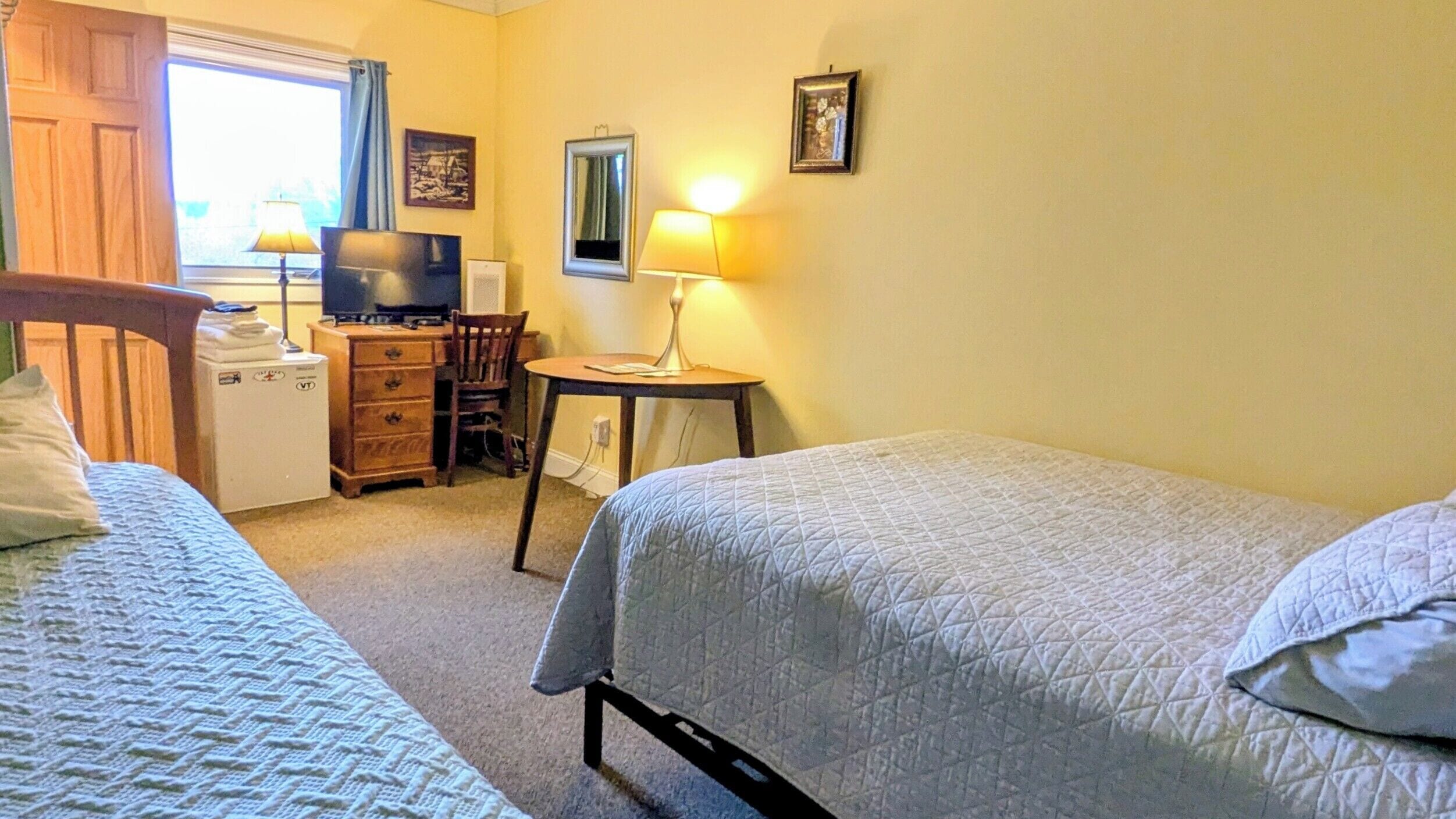 A bedroom with two beds, a desk with a chair, a television, and a lamp.