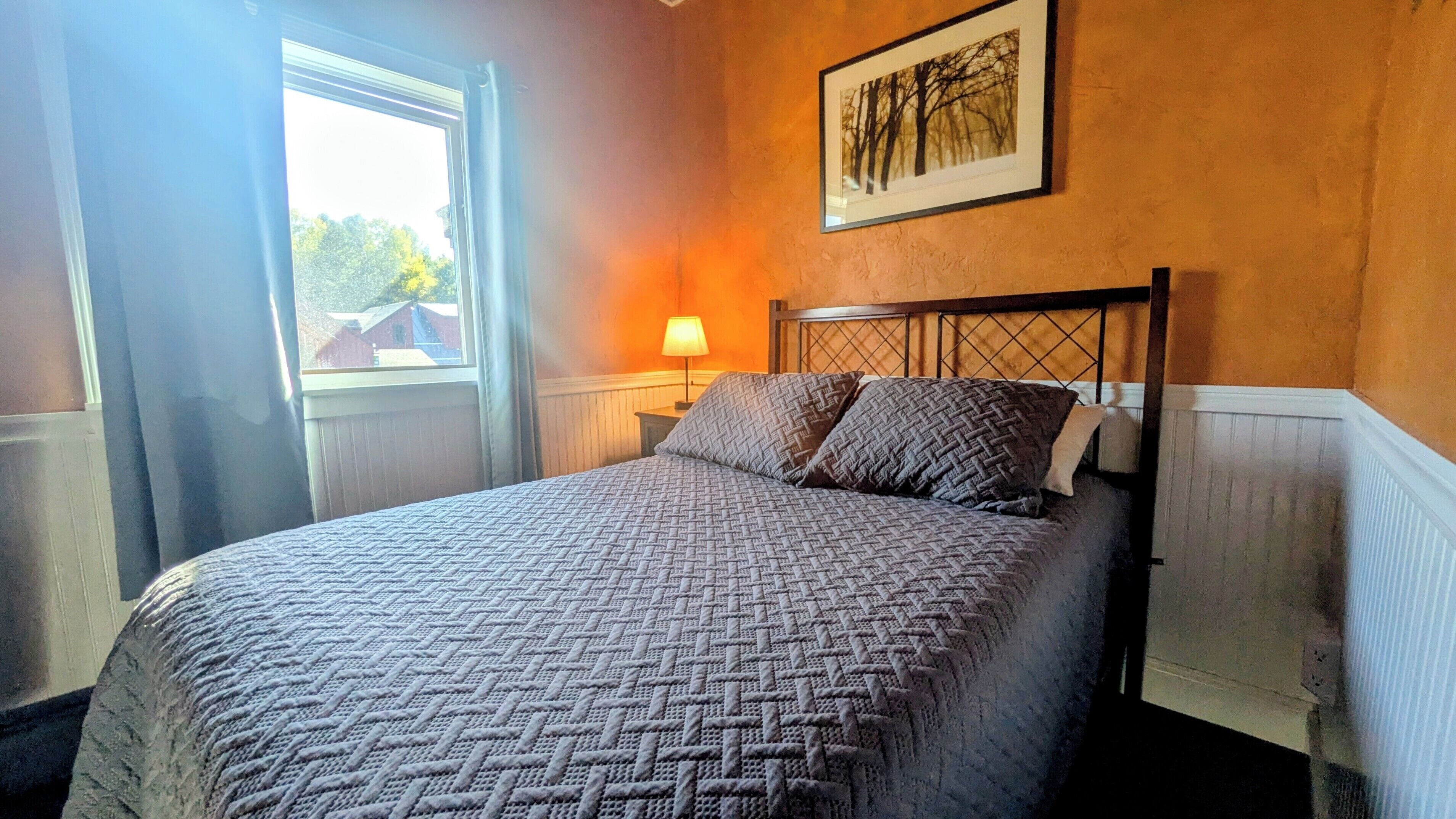Un dormitorio con cama, ventana, lámpara de pared y un cuadro enmarcado.