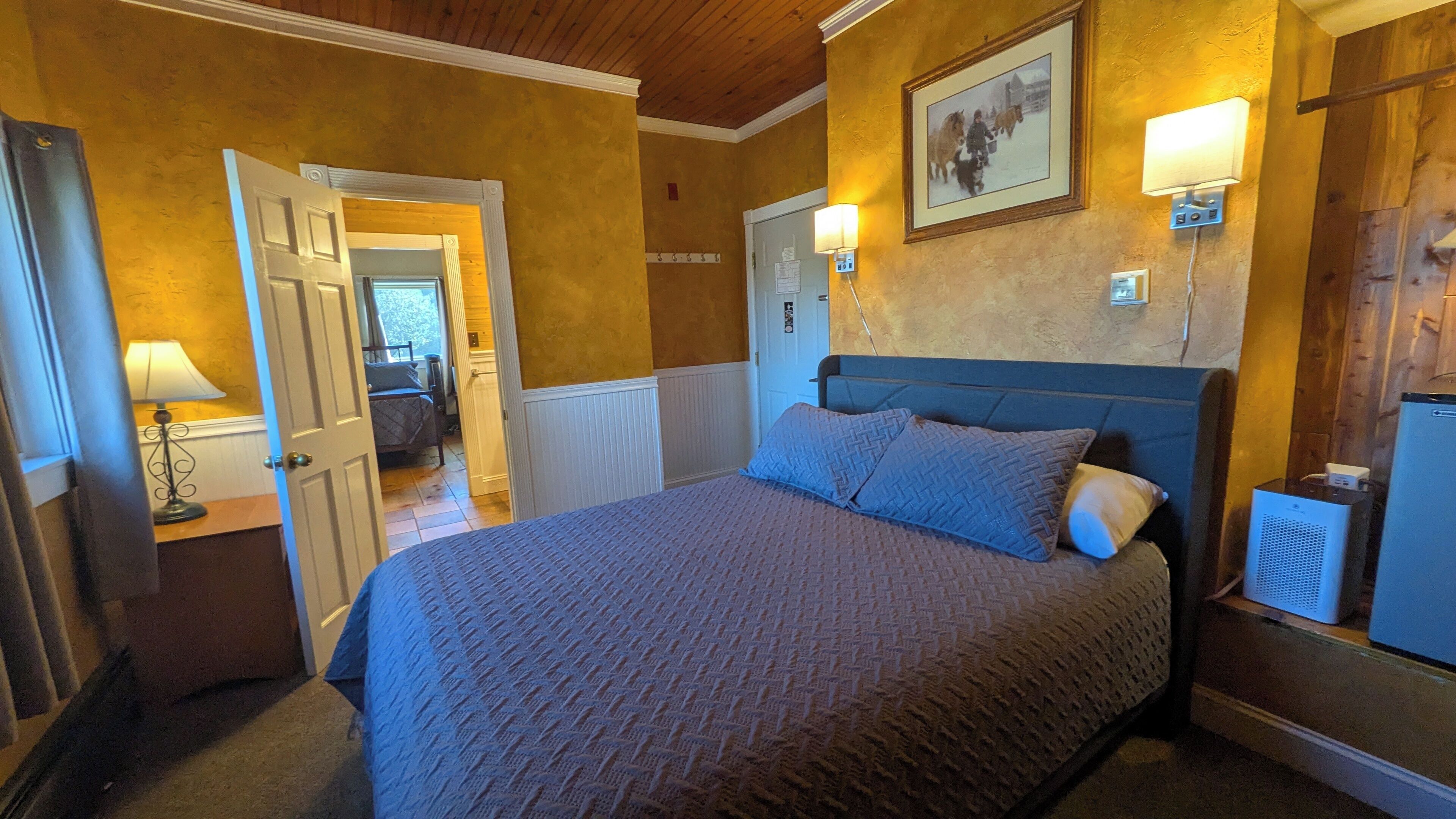 A bedroom with a large bed, a bedside table with a lamp, a wall-mounted heater, and a door leading to another room.