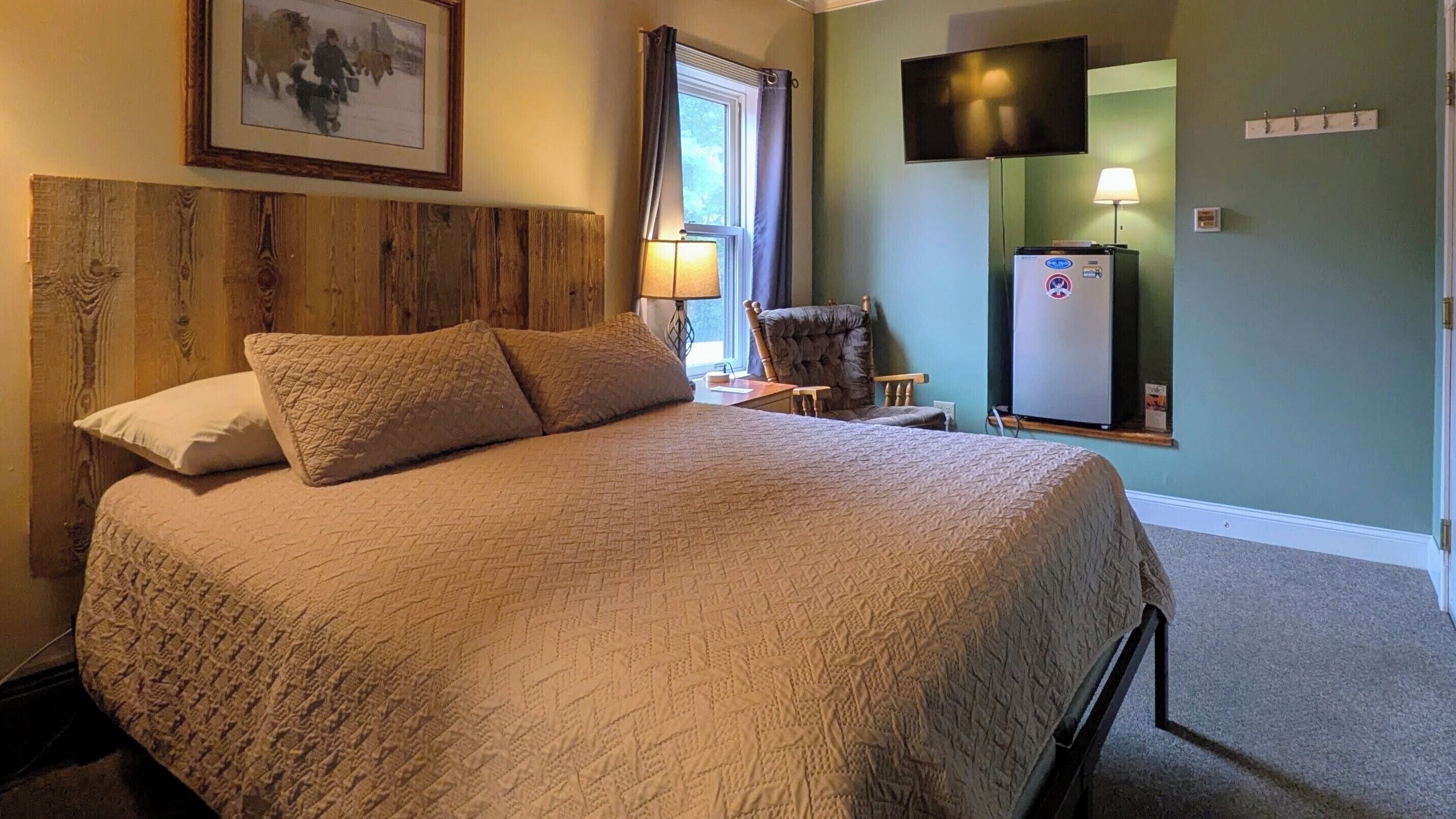 A neatly made bed with a quilted cover, a wooden headboard, a framed picture on the wall, a television mounted above a refrigerator, and a chair near the window.