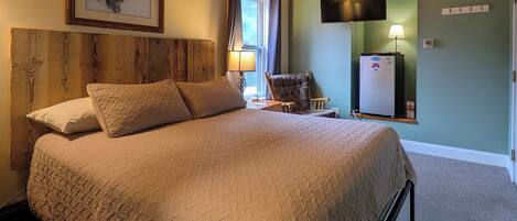 A neatly made bed with a quilted cover, a wooden headboard, a framed picture on the wall, a television mounted above a refrigerator, and a chair near the window.