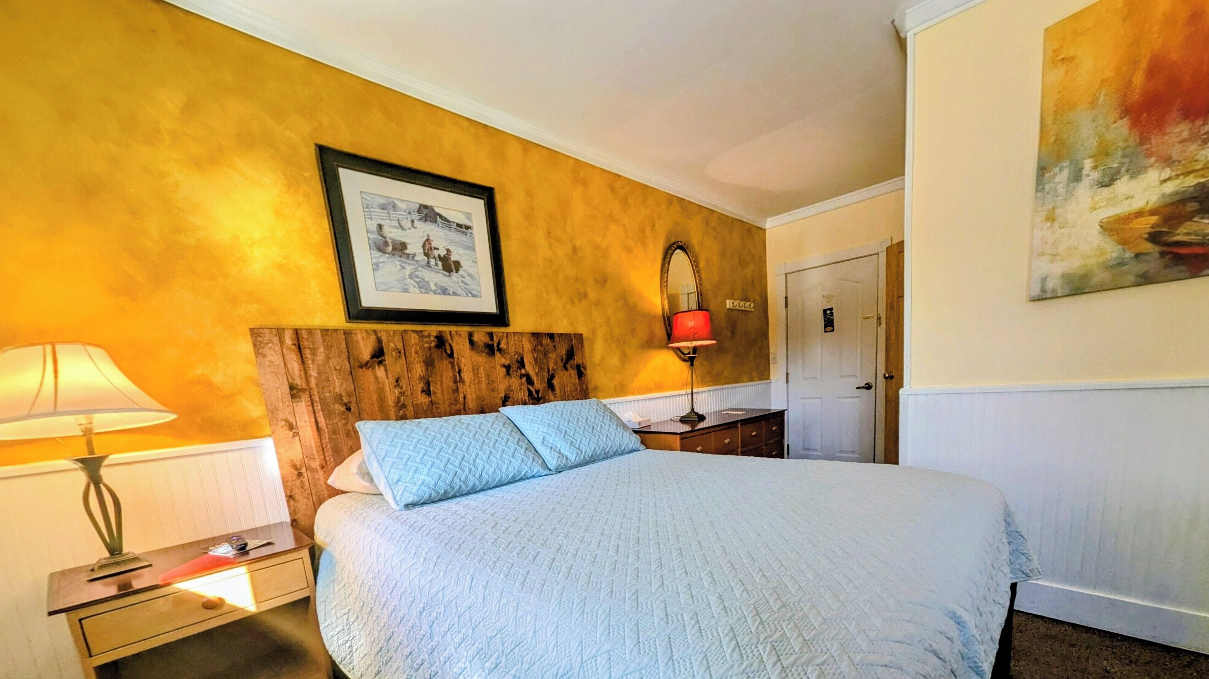 Een hotelkamer met een houten hoofdbord, een groot bed met een sprei, een nachtkastje met een lamp, een ingelijst schilderij aan de muur en een deur met een bordje.