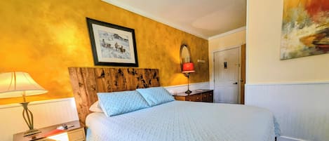 Een hotelkamer met een houten hoofdbord, een groot bed met een sprei, een nachtkastje met een lamp, een ingelijst schilderij aan de muur en een deur met een bordje.