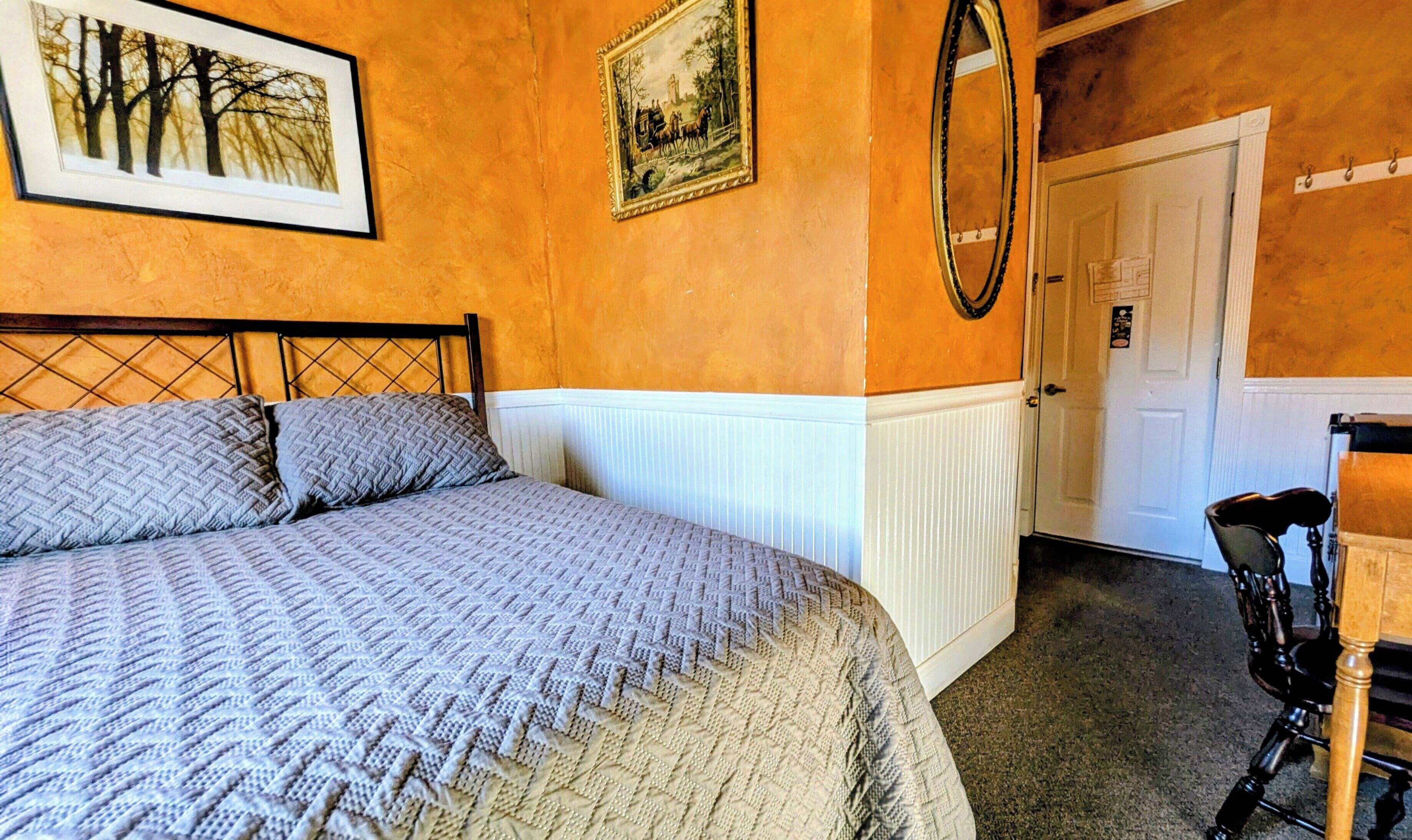 A bedroom with a bed, two pillows, a framed picture, a mirror, and a chair.