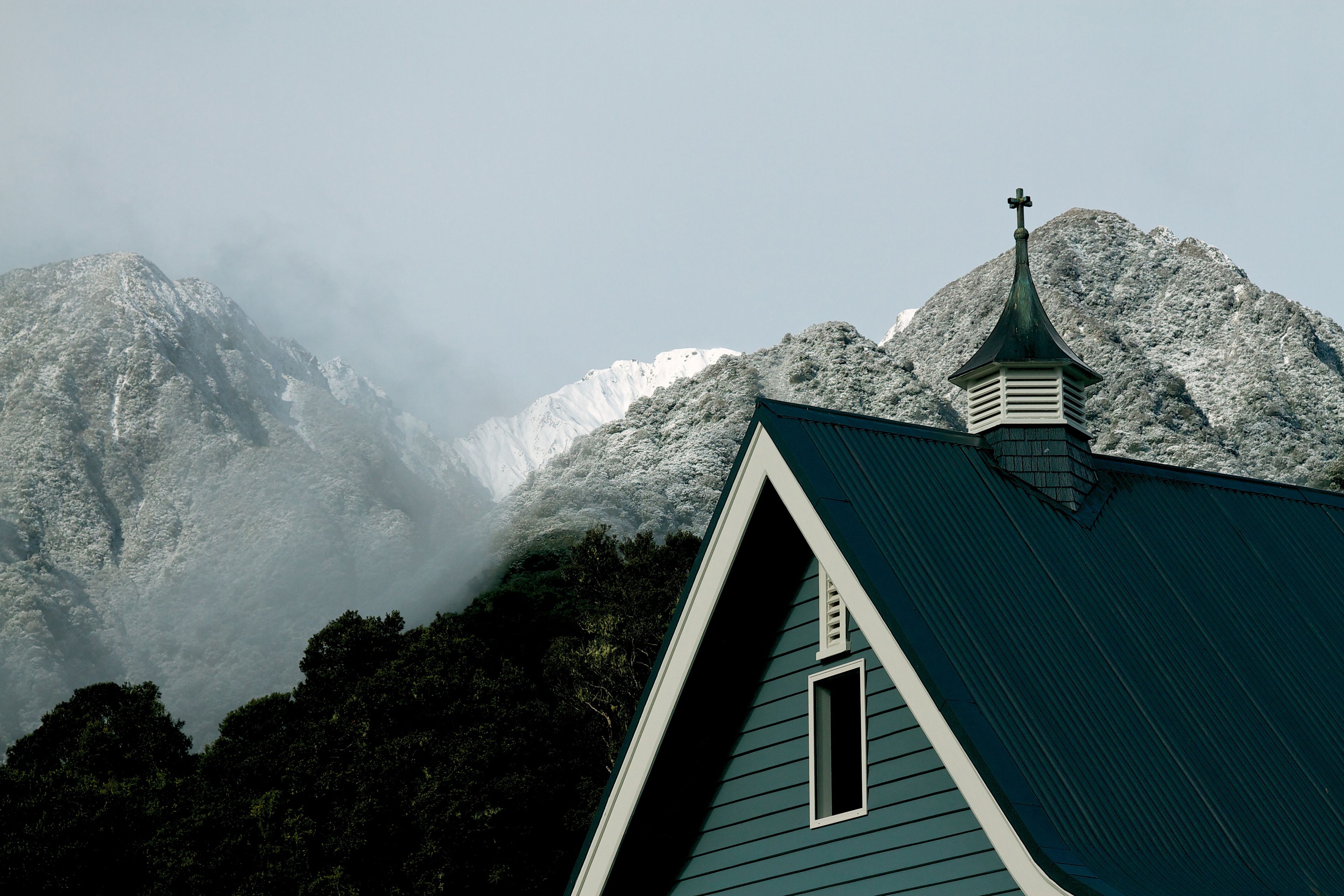 Uma igreja com telhado inclinado e uma cruz no topo, cercada por montanhas cobertas de neve e floresta densa.