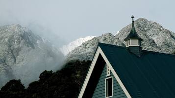 Uma igreja com telhado inclinado e uma cruz no topo, cercada por montanhas cobertas de neve e floresta densa.