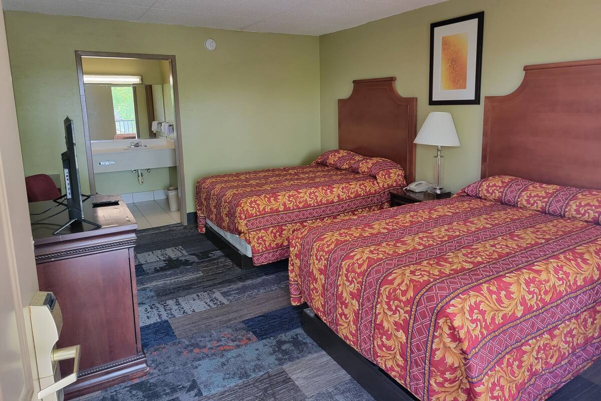 Ein Hotelzimmer mit zwei Betten, einem Schreibtisch mit Computer, einem Spiegel und einem durchgegangenen Türspalt einsehbaren Badezimmer.