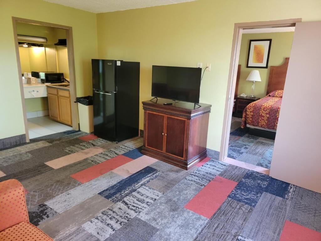 A hotel room with a kitchenette, a television on a wooden cabinet, a patterned carpet, and a bed with a patterned bedspread.
