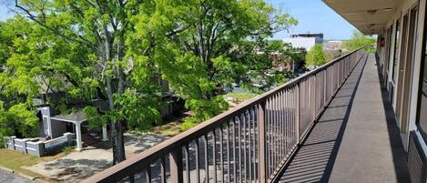 A covered outdoor corridor with a metal railing, trees, and a building in the background.