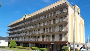 A multi-story hotel with balconies on each floor, a flat roof, and a parking area in front.