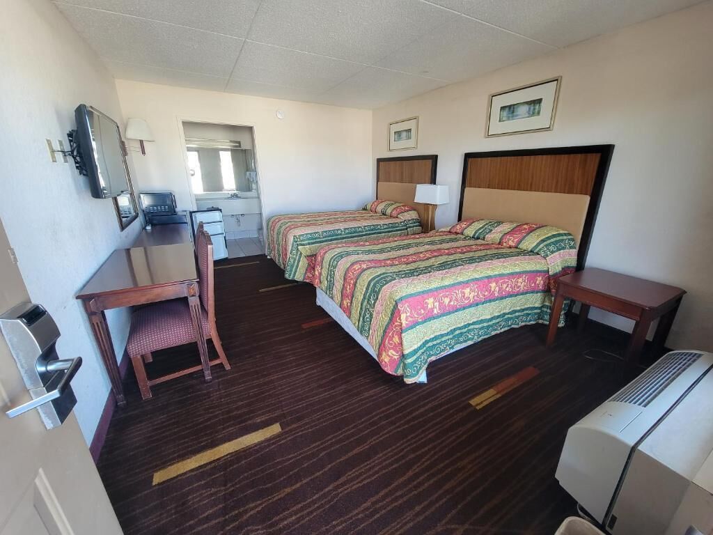 A hotel room with two beds, a desk, a chair, a mirror, a TV, and a heater.