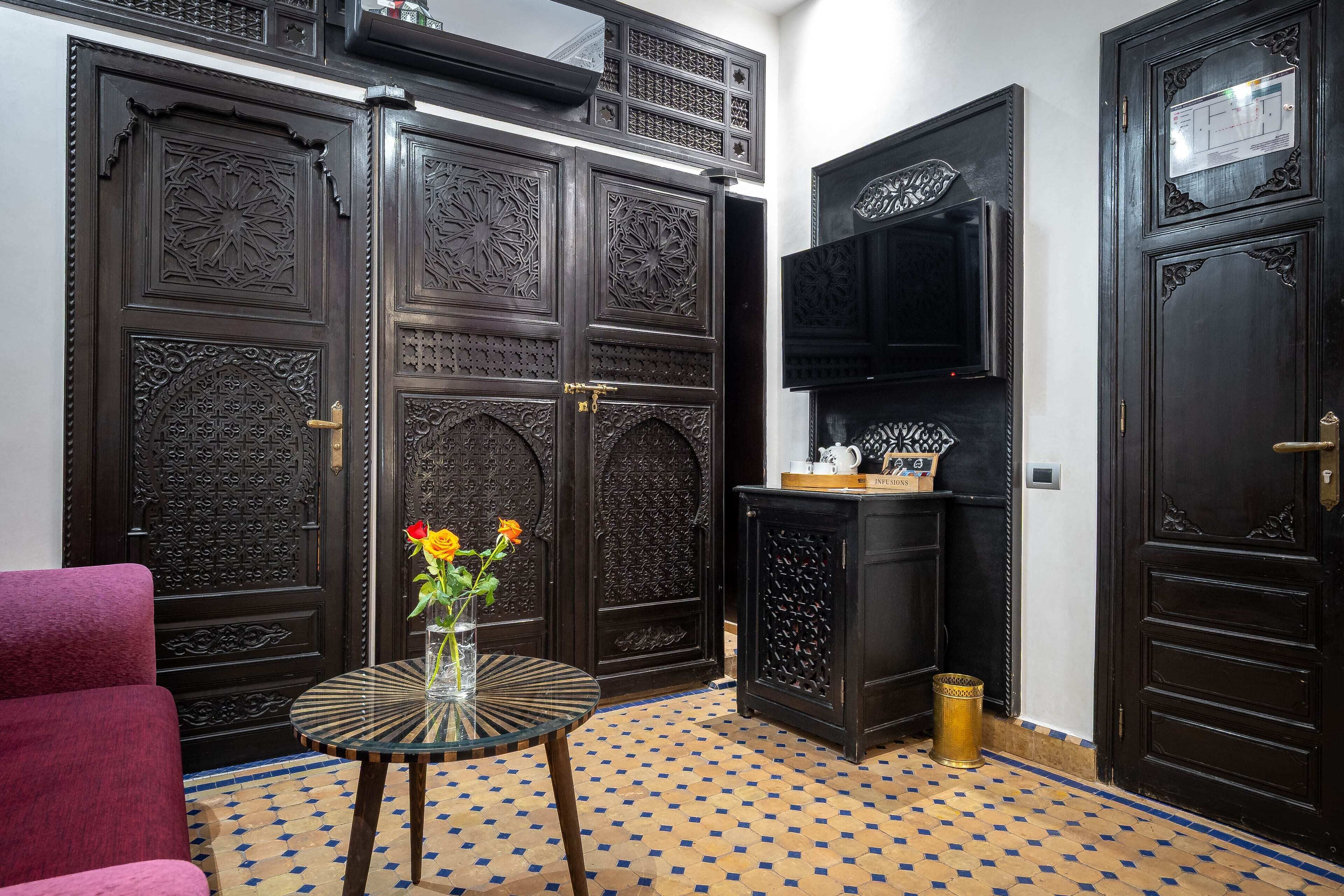 A room with ornate dark wooden doors, a flat-screen TV mounted on the wall, a small round table with a vase of flowers, and a patterned floor.
