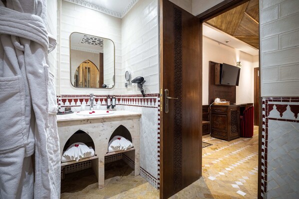 A bathroom with a white tiled wall, a mirror, a sink, a hairdryer, and a wooden cabinet. - Hotel & Ryad Art Place Marrakech (Marrakech)