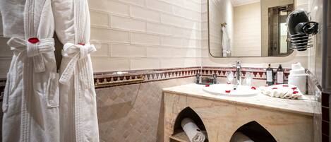 A bathroom with white tiled walls, a marble vanity, a mirror, and two white robes with red roses.