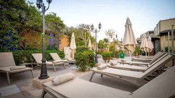 A sunlit outdoor lounge area with lounge chairs, umbrellas, and decorative street lamps.